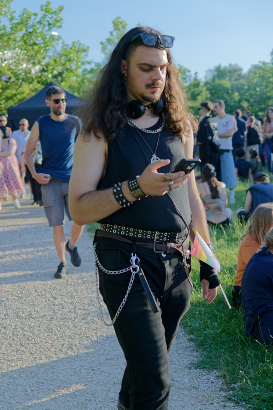 25 - Person wearing a black tank top and black pants, accessorized with layered silver necklaces and a studded belt, at a pride event